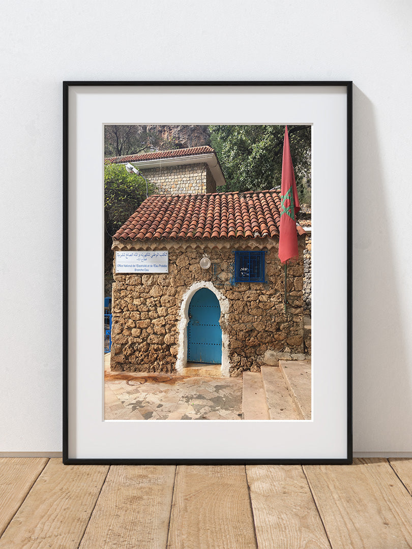 A photograph of a stone building with a blue door and red flag on a white wall in Chefchouen