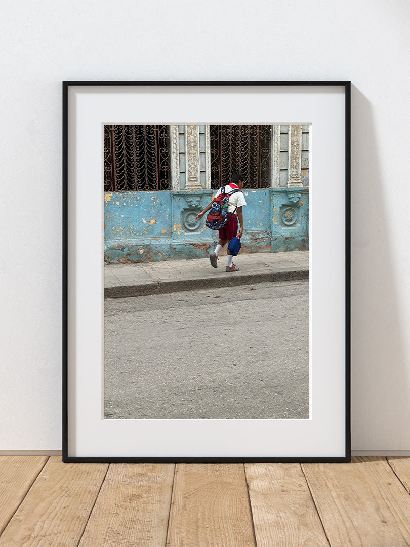 A photograph of a school boy walking on a Cuban street with a colorful building in the background.